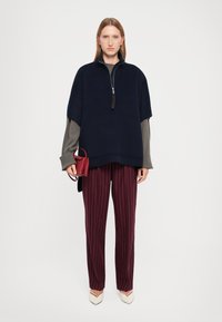 Pullover navy con colletto a zip, maniche lunghe grigie, pantaloni a righe bordeaux, e una piccola borsa rossa, completato da scarpe beige a punta.