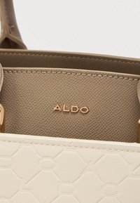 Balta rankinė su tekstūruotu paviršiumi, turinčia šešiakampių raštų. Viduje yra lygus taupe spalvos audinys su aukso spalvos "ALDO" ženklu. Matomi aukso akcentai.