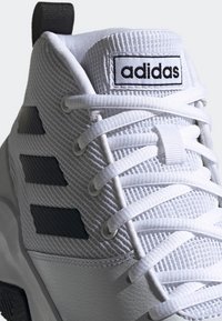 Baskets blanches Adidas avec des bandes noires, tige en mesh, languette rembourrée, lacets blancs et étiquette logo sur la languette.