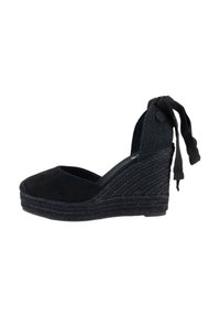 Sandalia cuña de esparto de ante negro con tacón de yute trenzado, punta abierta y una correa de tobillo tejida con lazos. Textura suave y diseño elegante.