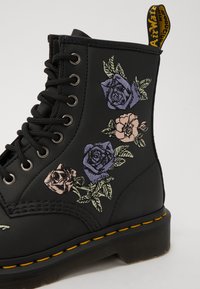 Svart läderankelstövel med broderade lila, rosa och gröna blommönster; har svarta snören och en gul dragflik bak.