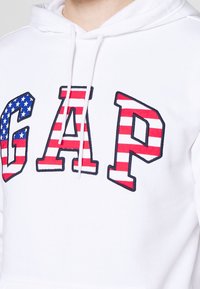 Sweat à capuche blanc avec de grandes lettres "GAP". Le "G" est bleu avec des étoiles, tandis que le "A" et le "P" sont rayés rouge et blanc. Cordon de serrage ajustable à la capuche.