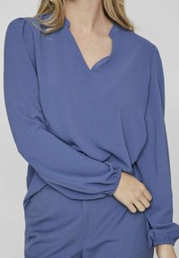 Modra bluzica z V-izrezom, ohlapnim krojem, teksturirano tkanino in elastičnimi manšetami. Rokavi imajo nežno nagubanje na ramenih.