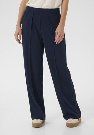 Mujer que lleva pantalones anchos azul oscuro y zapatillas beige, de pie con las manos relajadas a los lados sobre un fondo liso.