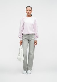 Lichtroze sweatshirt met pofmouwen met oogjesdetails, grijze wijde jeans, witte handtas en witte sneakers tegen een neutrale achtergrond.