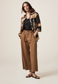 Braune weite Hosen mit Falten, kombiniert mit einem schwarzen Oberteil mit Spitze und einem gemusterten Oversized-Hemd in Erdtönen. Schwarze flache Sandalen.