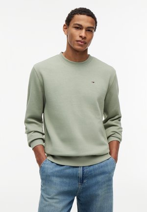 Noor mees, kes kannab helerohelist ümara kaelusega pusa ja siniseid teksaseid, käed taskutes, seisab ühevärvilise valge tausta ees.