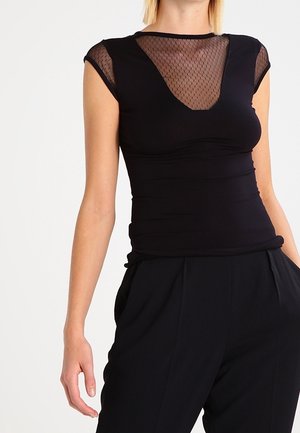 Kobieta ubrana w obcisły czarny top z przezroczystym siateczkowym dekoltem i krótkimi rękawami, w połączeniu z czarnymi spodniami z wysokim stanem na białym tle.