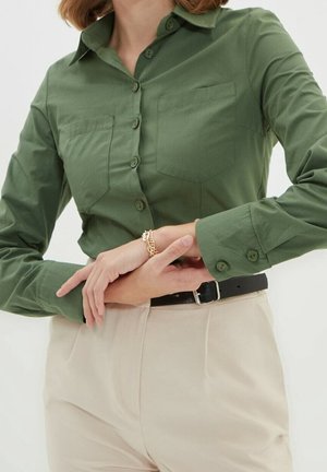 Chemise olive verte à boutons avec col pointu, deux poches poitrine, et manches longues avec poignets à boutons, associée à un pantalon de couleur claire.