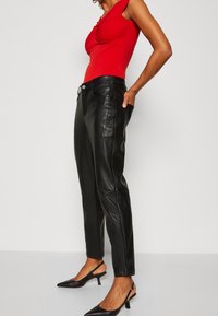 Červený přiléhavý top s uzlovým designem, zkombinovaný s šitými černými koženými kalhotami s bočními kapsami a zúženým kotníkem. Černé boty na podpatku.