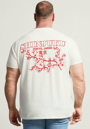 Homme portant un t-shirt blanc avec une branche de fleur de cerisier rouge et le texte « SHINJUKU TOKYO, JAPAN » dans le dos, associé à un jean bleu.