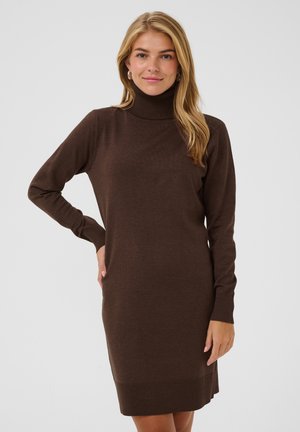 Femme portant une robe en maille marron à manches longues et col roulé, debout, une main sur la hanche, devant un fond gris clair uni.
