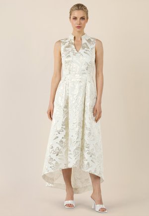 Frau trägt ärmelloses weißes High-Low-Kleid mit silbernem Blumenmuster und offenen weißen High Heels, steht vor schlichtem beigem Hintergrund.