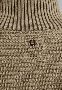Beige neulepaita, jossa on ribba korkea kaulus, ja joka sisältää tekstuuristymäkuvion sekä mustan ja oranssin logotagin ommeltuna kankaaseen.