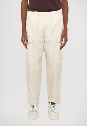 Beige cargo bukser med elastisk talje, der har to sidelommer og et let, krøllet materiale. Båret med hvide sneakers.