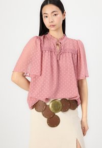 Rosa gepunktete Bluse mit Puffärmeln und Ausschnitt in Form eines Schlüssellochs, kombiniert mit einem braunen Leder-Gürtel mit kreisförmigen Bronze-Details.