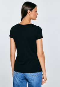 Kobieta o prostych ciemnych włosach, ubrana w czarną koszulkę z krótkim rękawem i niebieskie dżinsy, pokazana od tyłu na jednolitym tle.