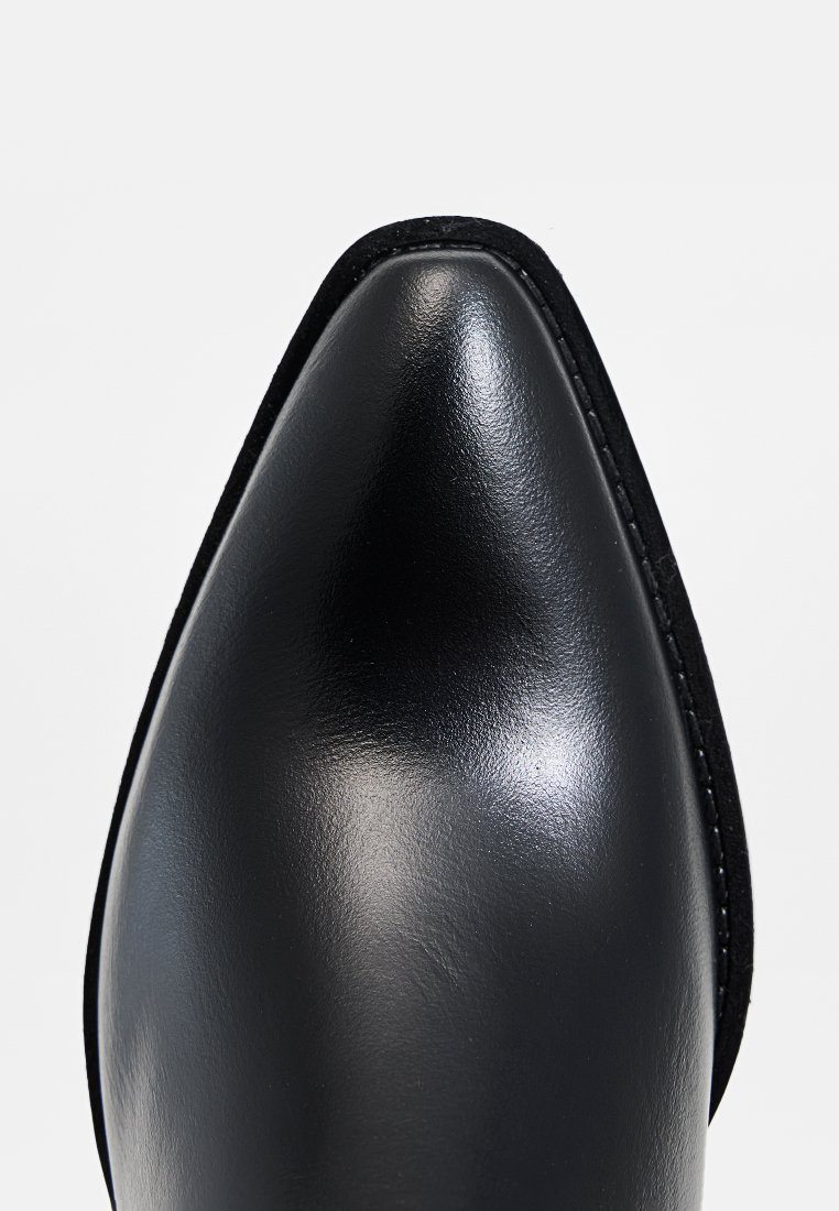 Punta de cuero negro brillante con acabado gloss, que presenta un contorno de ante negro contrastante y bordes cosidos de manera ordenada. Diseño simple y elegante.