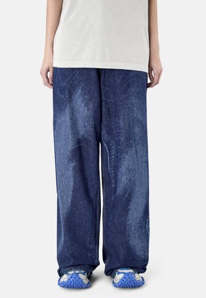 Osoba ubrana w luźne, szerokie jeansy z niebieskiego denimu, białą koszulę i białe trampki z niebieskimi, teksturowanymi podeszwami, stojąca na jednolitym tle.