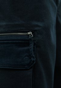 Μαύρο τζιν παντελόνι με φερμουάρ πλαϊνή τσέπη και μεταλλικά αξεσουάρ, σε υφασμάτινη υφή. Η κοντινή λήψη τονίζει τις λεπτομέρειες της τσέπης.