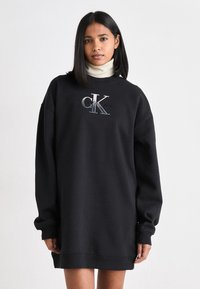Svart sweatshirt-klänning med långa ärmar, har rundad halsringning och broderad "CK"-logotyp i silver och blått. Mjuk tyg, avslappnad passform.