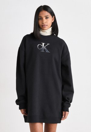 Rochie neagră din bumbac, cu mâneci lungi, având un gât rotund și un logo „CK” brodat în argintiu și albastru. Material moale, croială lejeră.