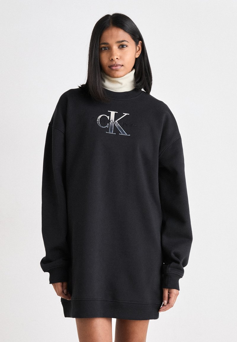 Svart sweatshirt-klänning med långa ärmar, har rundad halsringning och broderad "CK"-logotyp i silver och blått. Mjuk tyg, avslappnad passform.
