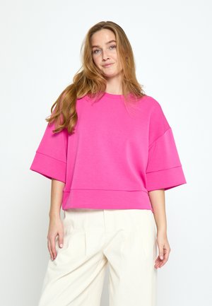 Jonge vrouw met lang haar die een felroze oversized top met korte mouwen en crèmekleurige broek met hoge taille draagt, staand tegen een witte achtergrond.