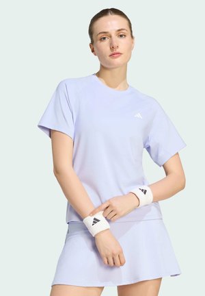Ung kvinde i lyse lilla atletisk trøje og nederdel, der justerer hvide armbånd med sort Adidas-logo, stående mod en bleg baggrund.
