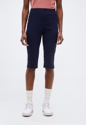 Személy térdig érő tengerészkék rövidnadrágot, fehér zoknit, fehér tornacipőt és rózsaszín felsőt visel, és egy egyszerű világos háttér előtt áll.