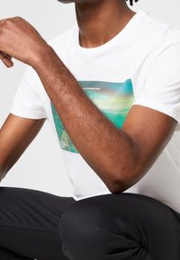 Camiseta de algodón blanca con un estampado gráfico colorido. Mangas cortas y un ajuste relajado. El estampado incluye tonos de verde y azul.