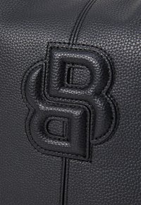 Musta nahkalaukku, jossa on teksturoitu pinta, kohokuvioitu logo ja erottuva ommel. Suunnittelussa on jämäkkä muoto ja huomattavia varusteita.