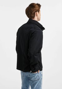 Czarna kurtka z wysokim kołnierzem, gładką fakturą i kieszeniami na zamek, zestawiona ze jasnoniebieskimi dżinsami, widok z tyłu.