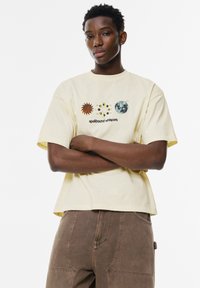 Korte mouwen gele T-shirt met een grafisch ontwerp van een zon, maan en planeten, en de tekst "betoverende fluisteringen." Gemaakt van zacht katoen.