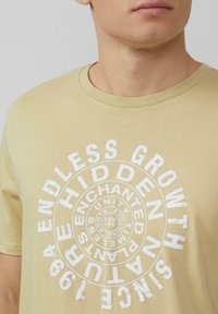 Zachte, lichtbeige T-shirt met ronde, witte grafische tekst met concentrische cirkels en verschillende woorden in vette letters. Katoen materiaal, korte mouwen.