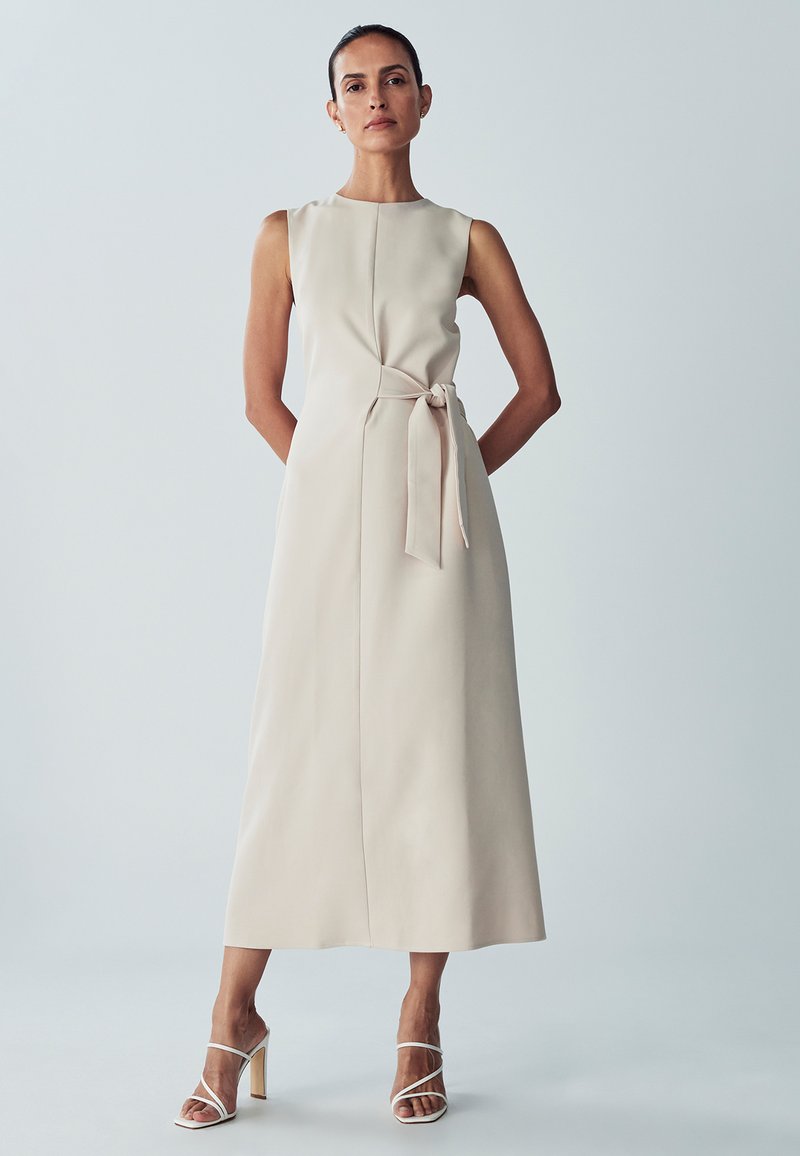 Beige mouwloze midi-jurk met een tailleband, met een rechte silhouette en zachte textuur. Gecombineerd met witte spitse hakken.