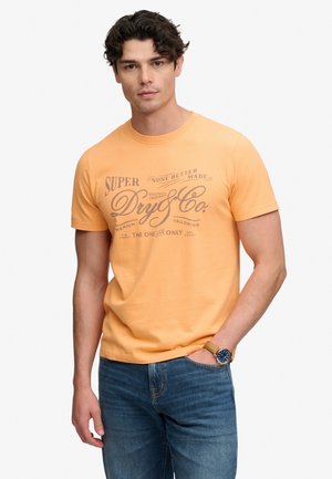 Uomo che indossa una t-shirt grafica pesca e jeans blu, con una mano in tasca e un orologio da polso con cinturino marrone.