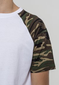 White cotton t-shirt with camouflage pattern short sleeves; features a round neckline and raglan sleeve design. Textured fabric accents.