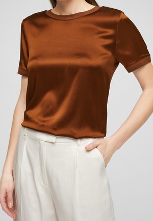 Frau trägt eine kurzärmelige braune Satinbluse, die in eine hoch taillierte weiße Hose mit Plisseefalten gesteckt ist, die Hand in der Tasche.