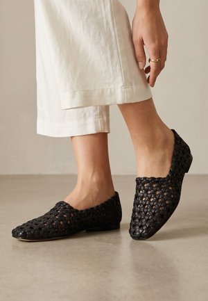 Persona con pantalones cortos color marfil y zapatos negros de tejido trenzado tipo slip-on, de pie sobre un suelo liso con la mano apoyada cerca del tobillo.