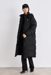 Cappotto lungo e nero in piumino con motivo trapuntato, cappuccio e chiusura con zip. Abbinato a un top nero aderente e a pantaloni neri, e un cappellino nero.