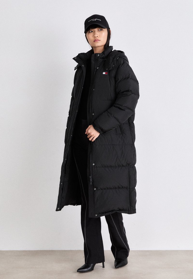 Schwarzer langer Daunenmantel mit Steppmuster, Kapuze und Reißverschluss. Kombiniert mit einem figurbetonten schwarzen Oberteil und einer schwarzen Hose sowie einer schwarzen Kappe.