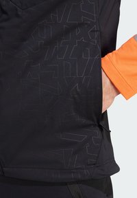 Schwarz atmungsaktives Sportshirt mit strukturiertem geometrischem Muster, ausgestattet mit einem orangefarbenen Ärmel und seitlichen Taschendetails für Funktionalität.