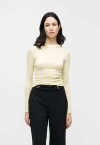 Απαλό κίτρινο μακρυμάνικο τοπ από μαλακό ύφασμα, συνδυασμένο με ψηλόμεσα μαύρα παντελόνια με κουμπιά για διακόσμηση. Ομαλή υφή συνολικά.