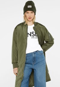 Olivgrüne Bomberjacke mit weichem Stoff, mit einem Reißverschluss vorne und gerippten Bündchen, kombiniert mit einem weißen Grafik-T-Shirt und blauen lässigen Jeans.