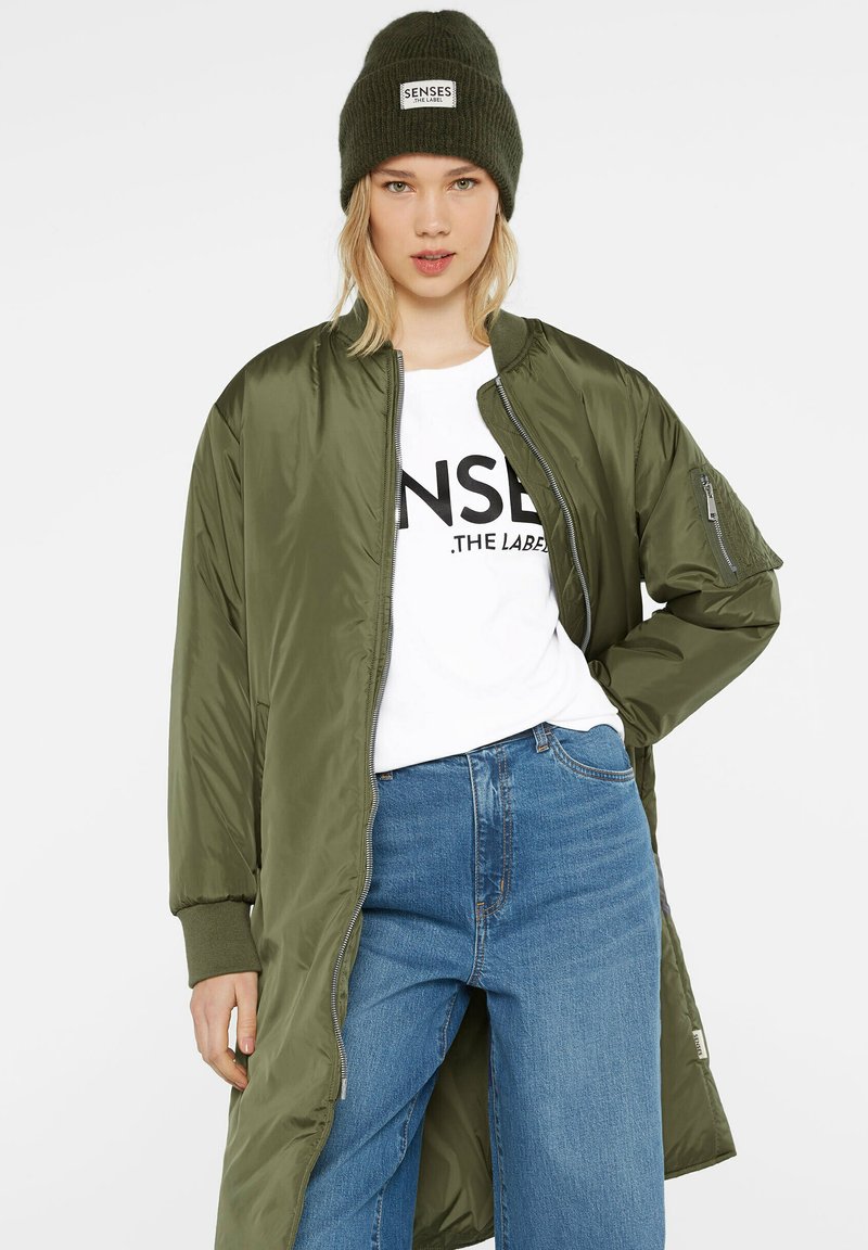 Olivgrüne Bomberjacke mit weichem Stoff, mit einem Reißverschluss vorne und gerippten Bündchen, kombiniert mit einem weißen Grafik-T-Shirt und blauen lässigen Jeans.