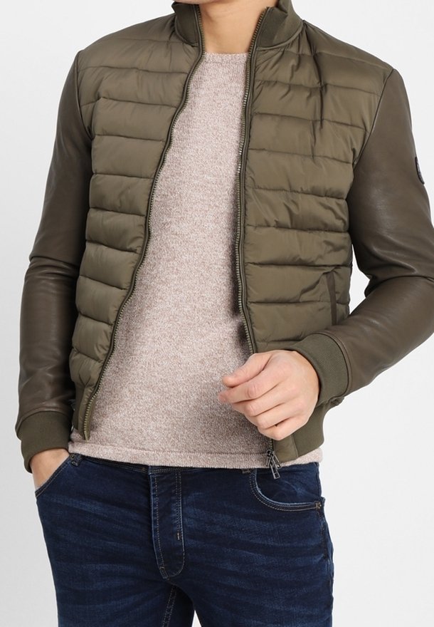 Man med olivgrön vadderad jacka med läderärmar, ljusbeige tröja och mörkblå jeans, stående med en hand i fickan.