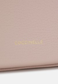 Light pink textured leather bag with gold "Coccinelle" logo, featuring smooth edges and subtle grain pattern.
