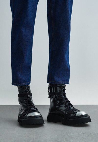 Persona che indossa stivali da combattimento in pelle nera con punte metalliche e lacci neri, abbinati a jeans dritti blu scuro su un pavimento grigio.