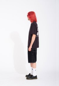 Svart t-shirt med grafisk design, svarta denimshorts, vita strumpor och svarta skor. Modellen står vänd mot vänster mot en vit bakgrund.